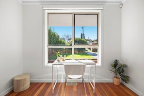 A room with a white desk, chair, and a large window with a view of a garden and houses. - Domi Rentals - The Maggie Home (Clayton)