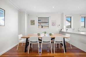 A modern dining area with a wooden table and white chairs, a window with a view, and a white tiled backsplash. - Domi Rentals - The Maggie Home (Clayton)