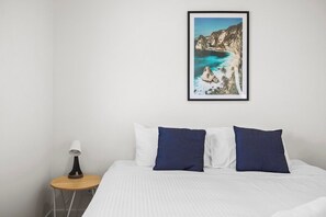 A bedroom with a bed, pillows, a nightstand, a lamp, and a framed picture of a coastal landscape. - Domi Rentals - The Maggie Home (Clayton)