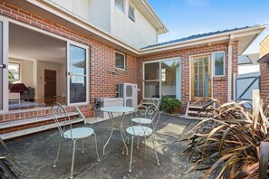 A patio with a brick wall, a sliding glass door, and a metal table and chairs. - Domi Rentals - The Maggie Home (Clayton)