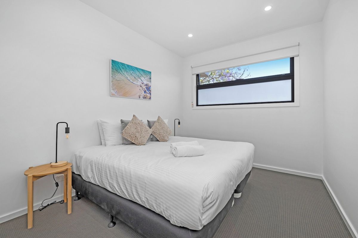 Un dormitorio con cama, mesita de noche, lámpara, ventana y un cuadro en la pared.
