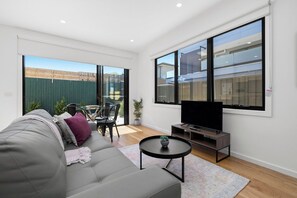 A modern living room with a grey sofa, a flat-screen TV, a small round coffee table, and a large window offering a view of the outside.