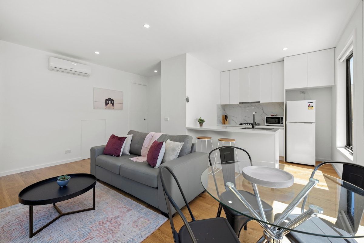 A modern living space with a grey sofa, a glass dining table, and a kitchen area with white cabinets and a marble countertop.