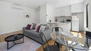 Ein zeitgemäßer Wohnbereich mit grauem Sofa, einem Glastisch und einer Küchenzeile mit weißen Schränken.
