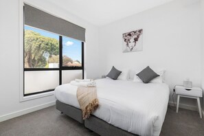 A bedroom with a large window, a bed with white bedding, a gray headboard, and a bedside table with a lamp.