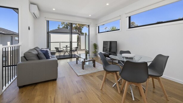 A modern living room with a sofa, dining table, chairs, a flat-screen TV, and large windows.