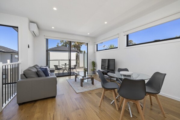 A modern living room with a sofa, dining table, chairs, a flat-screen TV, and large windows.