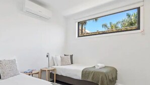 A bedroom with a bed, a bedside table, a window with a view of palm trees, and an air conditioning unit.