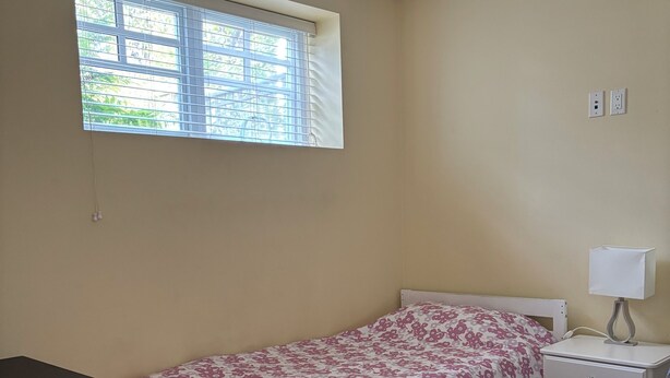 Una cama individual con colcha floral, una mesita de noche con lámpara, una ventana con persianas y un enchufe en la pared.