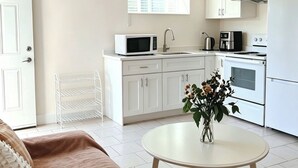 Una cocina moderna con armarios blancos, un microondas, una cafetera y un horno. Hay una silla blanca y una mesa redonda con un florero.