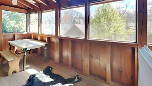 Una habitación con paredes de madera, ventilador de techo, una banca, una mesa y ventanas amplias con vista a árboles.