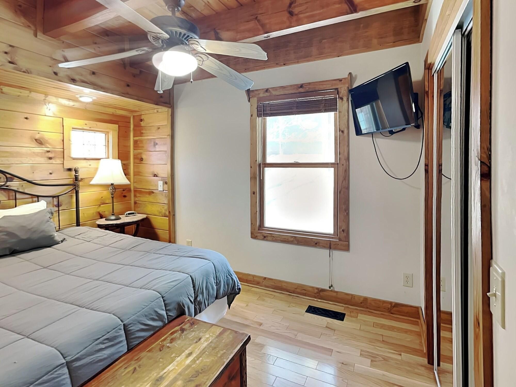 A bedroom with a bed, a nightstand, a lamp, a ceiling fan, a TV mounted on the wall, and a window.