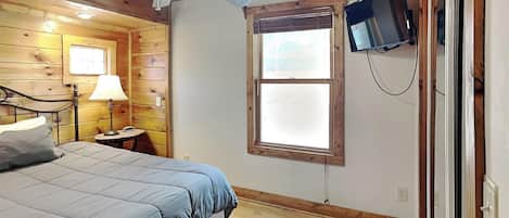 A bedroom with a bed, a nightstand, a lamp, a ceiling fan, a TV mounted on the wall, and a window.