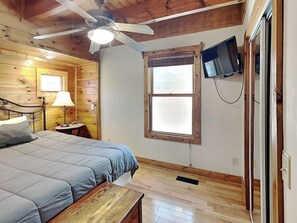 A bedroom with a bed, a nightstand, a lamp, a ceiling fan, a TV mounted on the wall, and a window. - Smokey Bear Haven (Pigeon Forge)