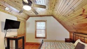 Un dormitorio con techo inclinado, una cama con ropa de cama floral, un televisor sobre una mesa de madera y una ventana con persianas.