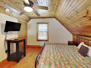 A bedroom with a sloped ceiling, a bed with floral bedding, a television on a wooden table, and a window with blinds.