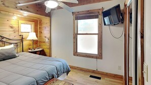 Una habitación con cama, escritorio, ventilador de techo, ventana y televisor montado en la pared.
