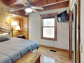 A room with a bed, a desk, a ceiling fan, a window, and a TV mounted on the wall.