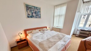 A bedroom with a large bed, a bedside table with a lamp, a radiator, a window with blinds, and a painting on the wall. - Hotel Zlatvo K Ov (Teplice)