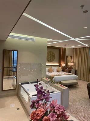 A hotel room with a large bed, a sitting area with a desk, a mirror, and a view of the city. - Rio Hotel (Riyadh)