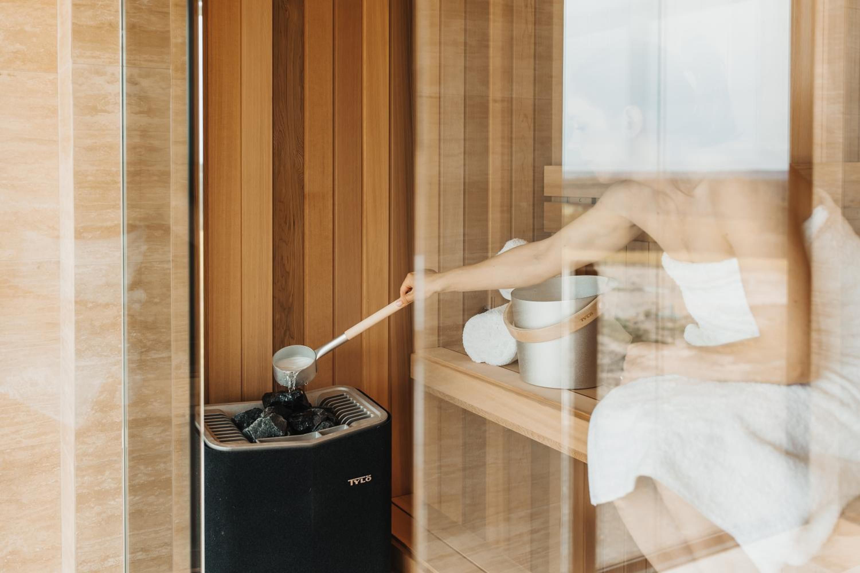 A sauna with a wooden interior, a black sauna heater, and a person using a ladle.
