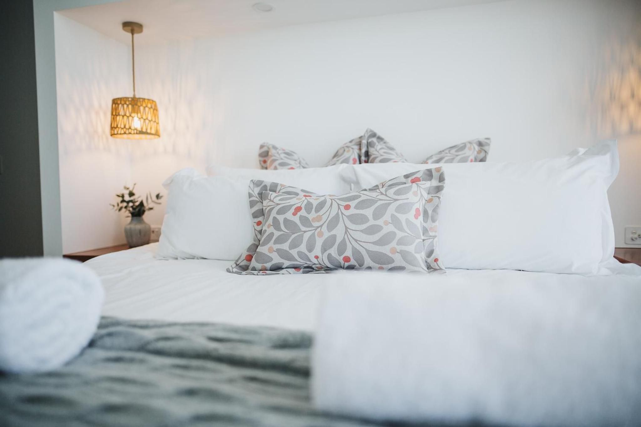 A neatly made bed with white bedding and patterned pillows, a bedside lamp, and a vase with flowers on a nightstand.