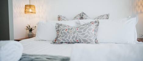A neatly made bed with white bedding and patterned pillows, a bedside lamp, and a vase with flowers on a nightstand.