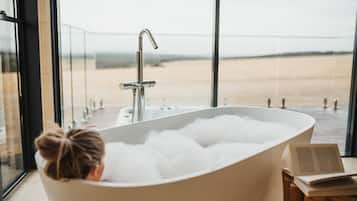 A bathtub filled with bubbles, a window with a view of a beach, and a book on a wooden surface.