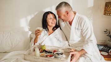 A couple in a hotel room with a breakfast tray containing fruits, yogurt, and a drink.