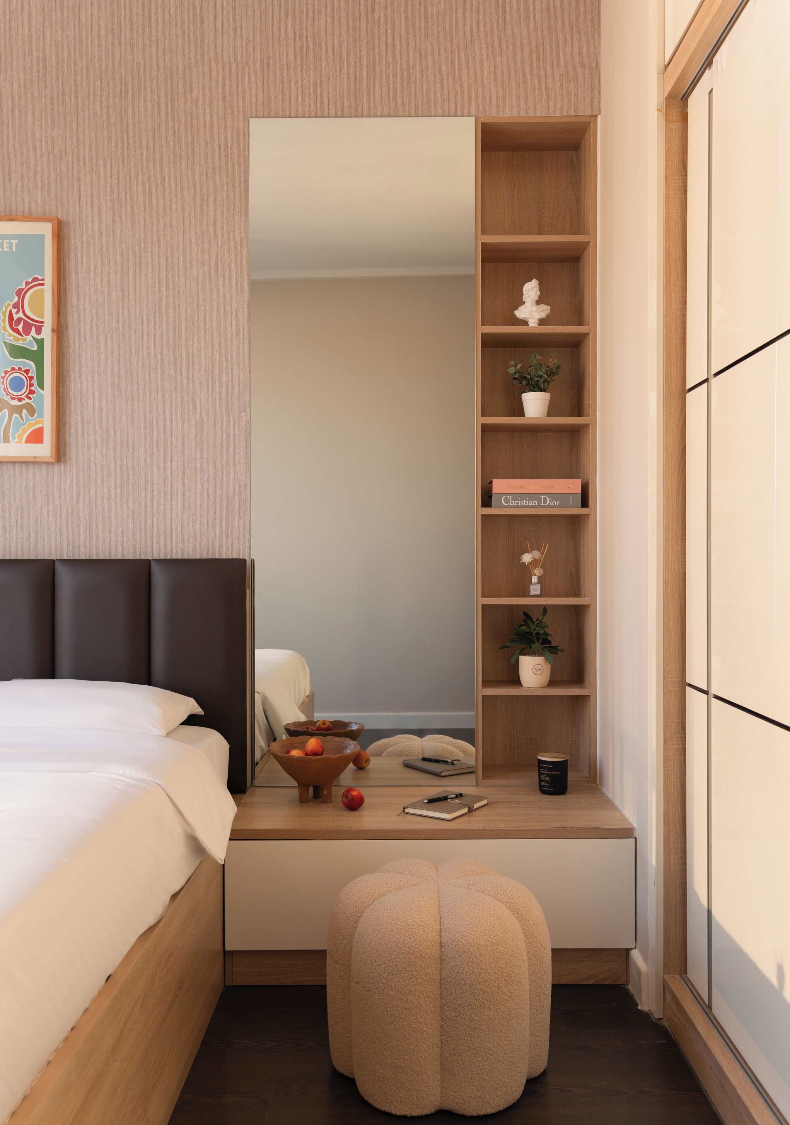 A bedroom with a bed, a bedside table with a mirror, a shelf with books and decorative items, and a small stool.