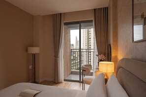 A hotel room with a bed, a chair, a lamp, a view of buildings through a window, and a balcony. - Maison De Gu Rivergate (Ho Chi Minh City)