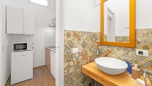 Ein kleines Badezimmer mit Waschbecken in Weiß, einem Spiegel mit Holzrahmen, einer Mikrowelle und einem kleinen Schrank.