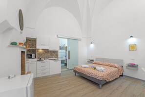 A bedroom with a bed, a small bedside table, a wall-mounted light, a microwave, and a kitchen area with cabinets and a sink. - Casa Miggiano 6 BR Apartment in Town, Santa Cesarea Terme, Italy (Santa Cesarea Terme)