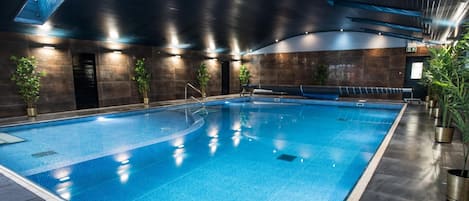 Una amplia piscina interior con suelo de baldosas, rodeada de paredes oscuras y plantas en macetas.