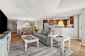 A spacious living room with a gray sofa, a coffee table, and a dining area with chairs and a table. The kitchen is visible in the background with wooden cabinets and a refrigerator. - Gorgeous 5th Floor Oceanfront Views | Pet Friendly | Super Spacious 1 Bedroom (Fernandina Beach)