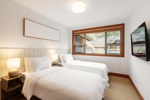 A bedroom with two beds, a nightstand, a lamp, a window with a view of buildings, and a framed picture on the wall. - Modern 3BR | Walk to Whistler Village | Hot Tub | AC | Pet-Friendly (Whistler)