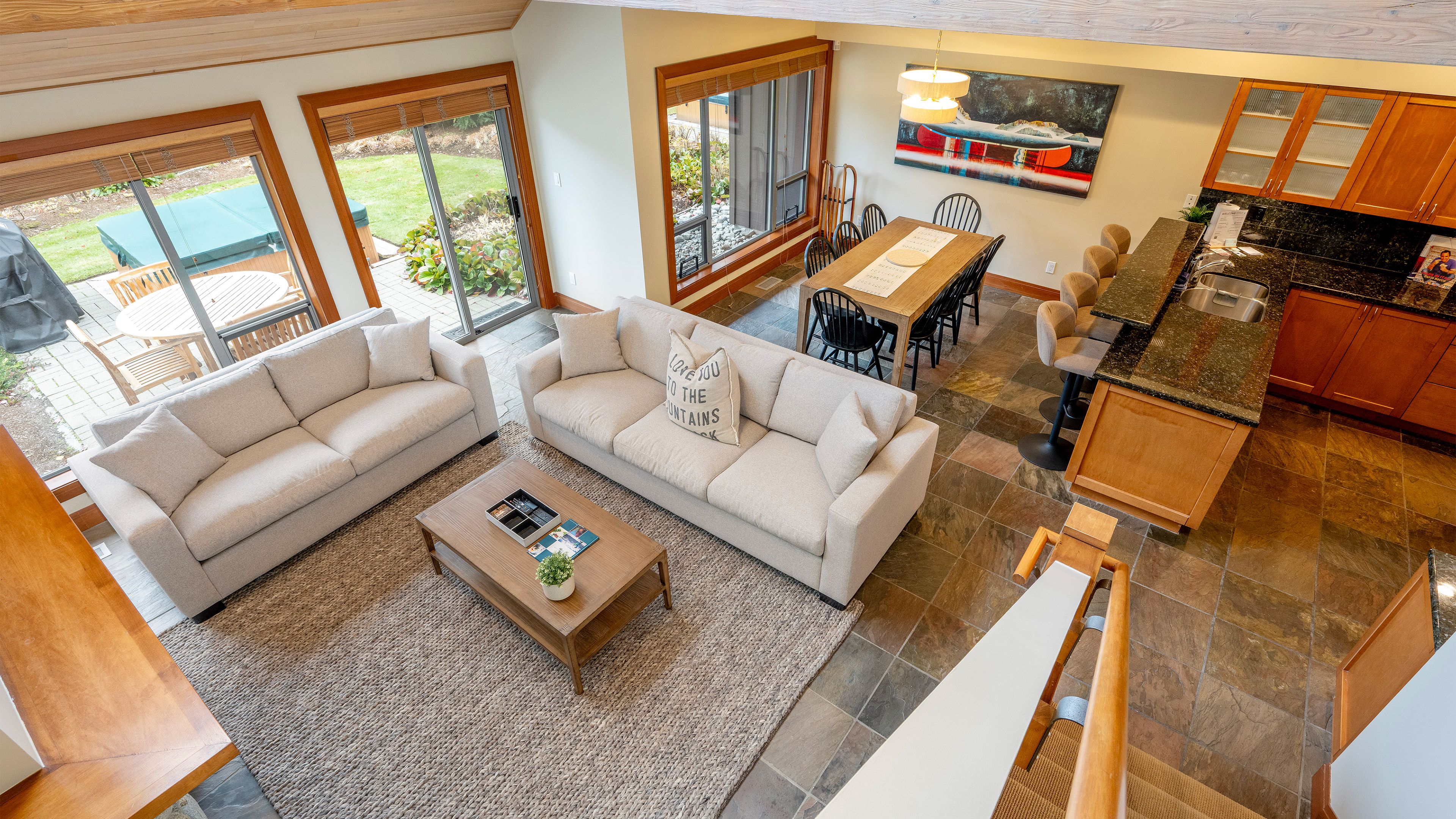 A spacious living area with a large sofa, a coffee table, and a dining table. There are large windows providing views of the outdoors.
