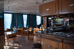 Un restaurante con mobiliario de madera, una zona de bar y grandes ventanales con cortinas.