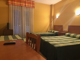 Habitación de hotel con dos camas, una mesa de madera, un televisor sobre un mueble bajo y una ventana con cortinas.