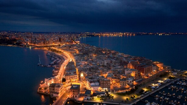 Vista aerea di una città costiera con un porto turistico, edifici e una strada lungo il lungomare.
