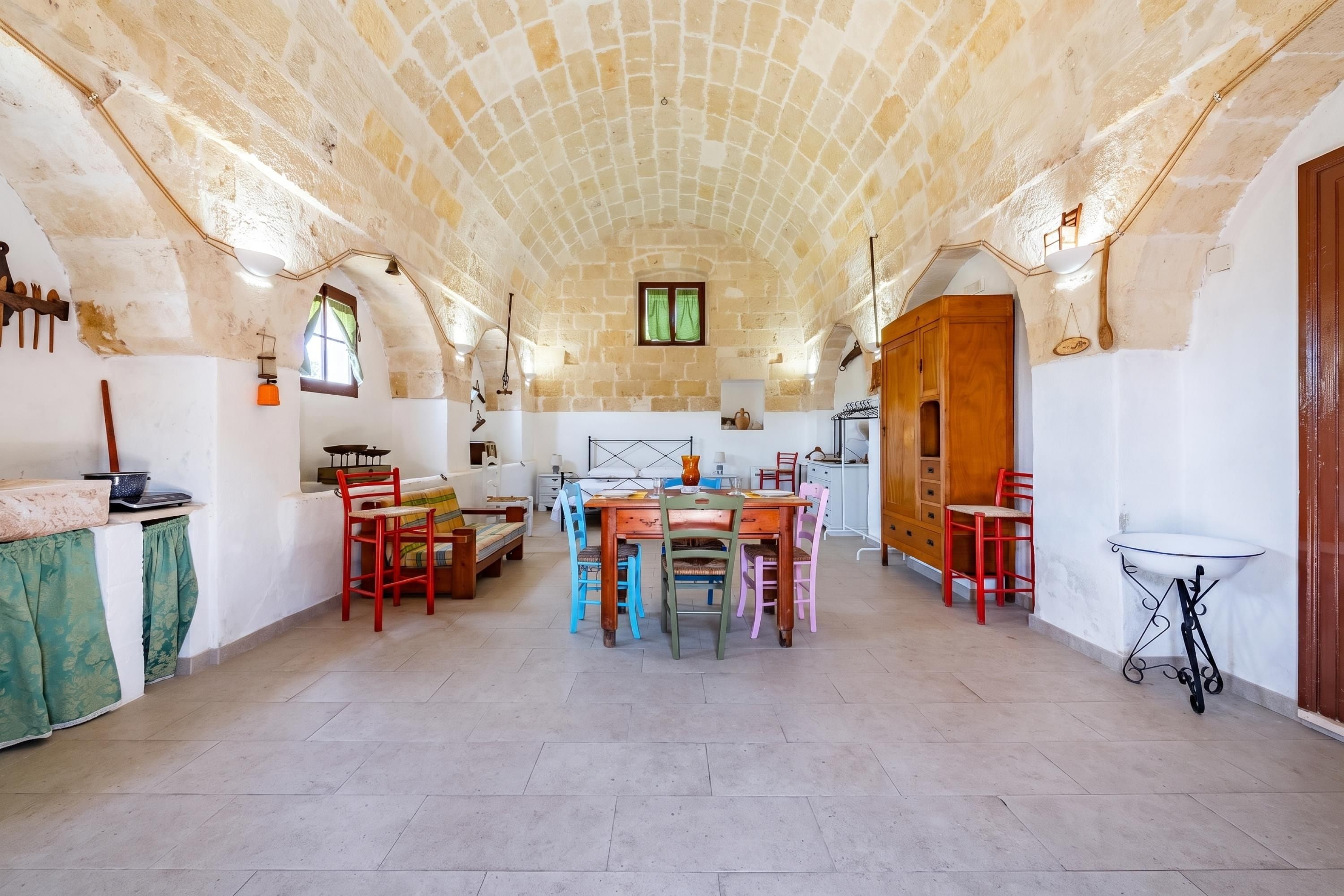 A spacious room with a high arched ceiling, a dining area with colorful chairs, a kitchen with wooden cabinets, and a small table with a white top.