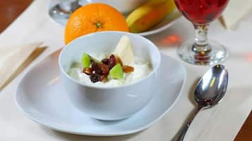 Una mesa con mantel blanco, un tazón de yogur con frutas y nueces, un vaso de jugo de naranja, una copa de vino tinto y un plátano.