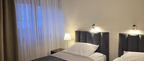 A hotel room with two beds, a nightstand, a lamp, and a window with curtains.