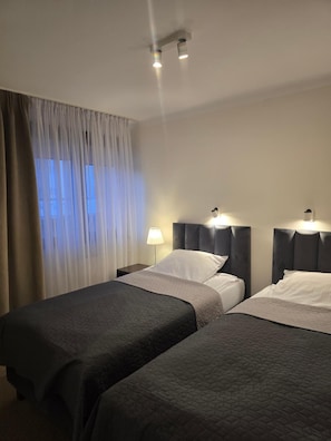 Habitación de hotel con dos camas, una mesita de noche, una lámpara y un ventanal con cortinas.