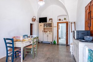 A dining area with a table set for four, chairs, a television above the table, and a kitchen area with cabinets and a stove. - Masseria Le Mesolecchie - Salvia Apt, Crispiano, Italy (Crispiano)