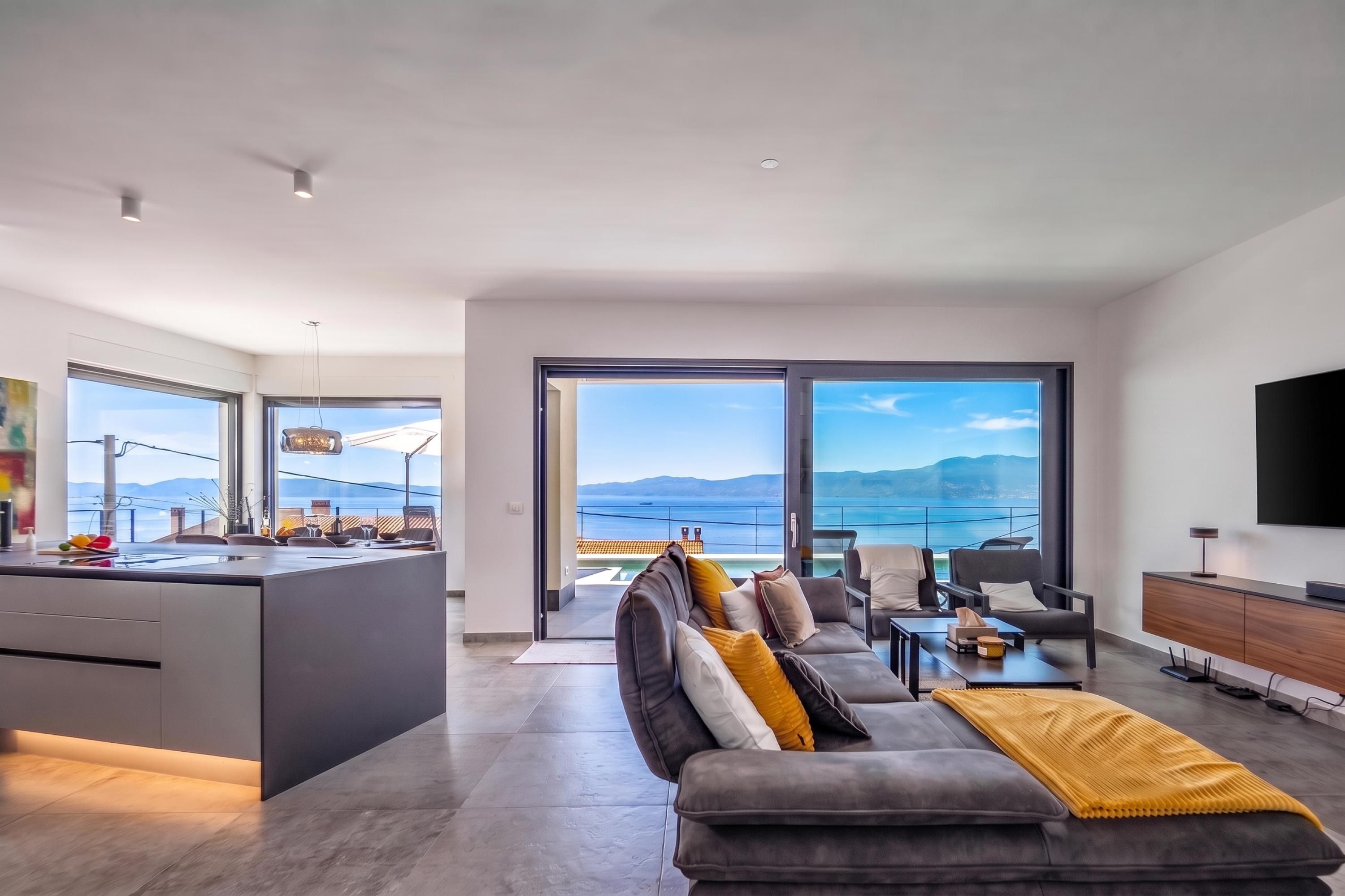 A modern living room with a large sofa, a coffee table, and a TV. There is a kitchen area with a bar and a view of the sea.