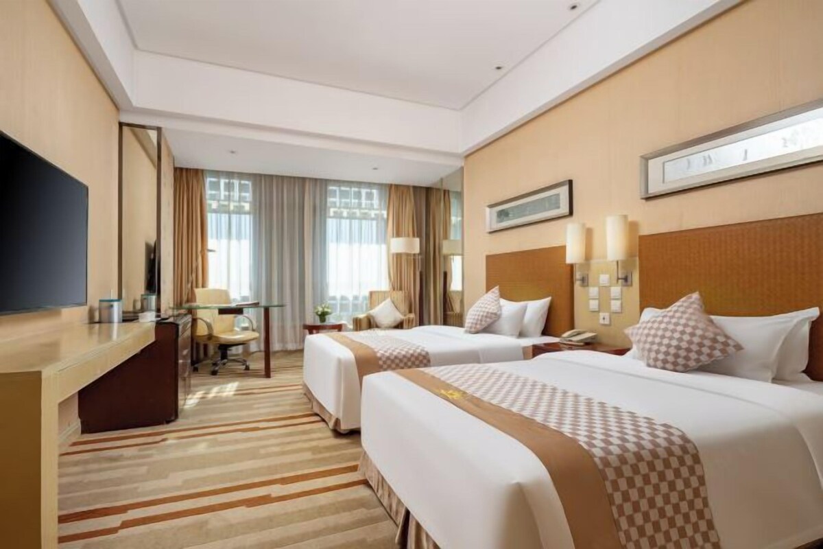 A hotel room with two beds, a desk, a TV, and a large window with curtains.