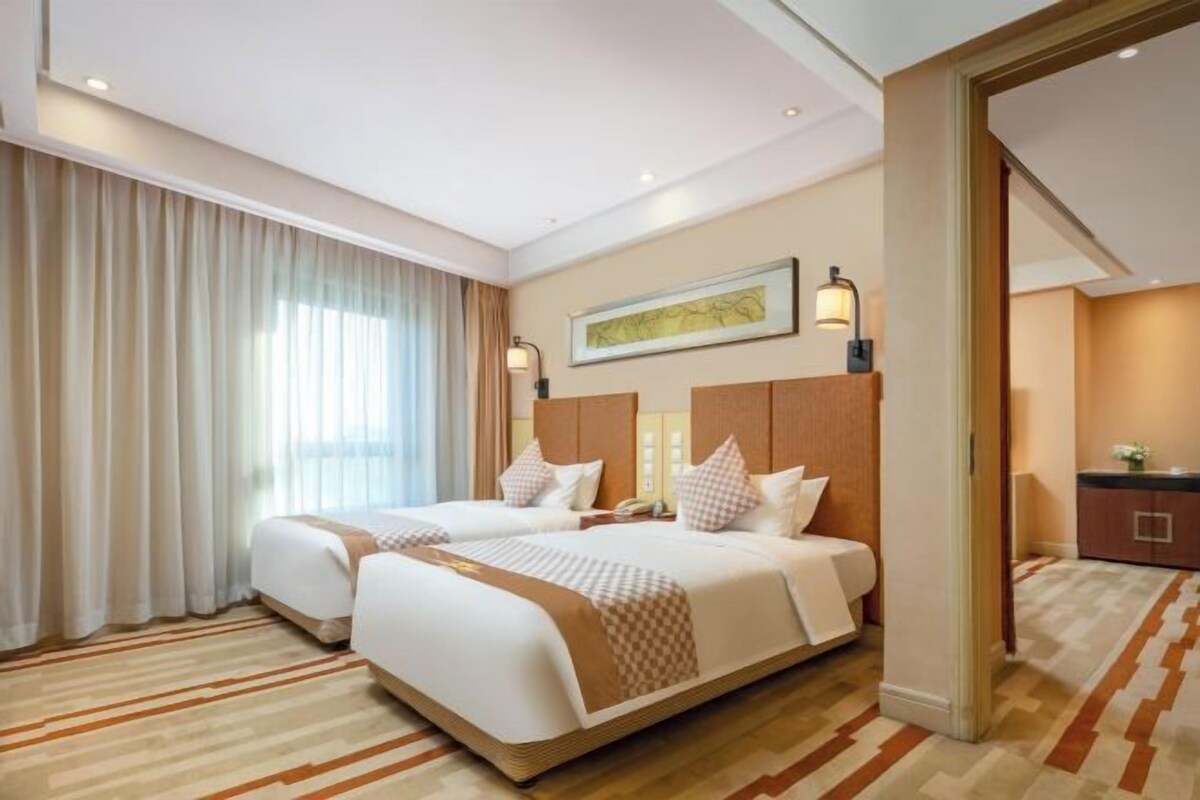 A hotel room with two beds, a large window with curtains, a painting on the wall, and a carpeted floor.