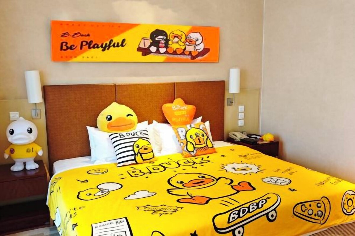 A hotel room with a bed featuring a yellow duck-themed bedspread, a wall-mounted orange sign with cartoon ducks, and a plush duck toy on the nightstand.