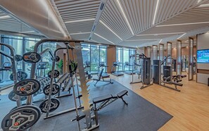 Academia moderna com diversos equipamentos de exercício, grandes janelas e piso de madeira.